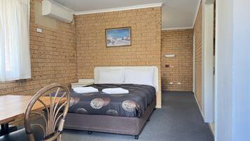 Habitación de hotel pequeña con pared de ladrillo, una cama individual con colcha estampada, una mesita con sillas y una ventana con cortinas.