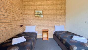 A room with two single beds, a small wooden nightstand, a painting on the brick wall, and a carpeted floor.