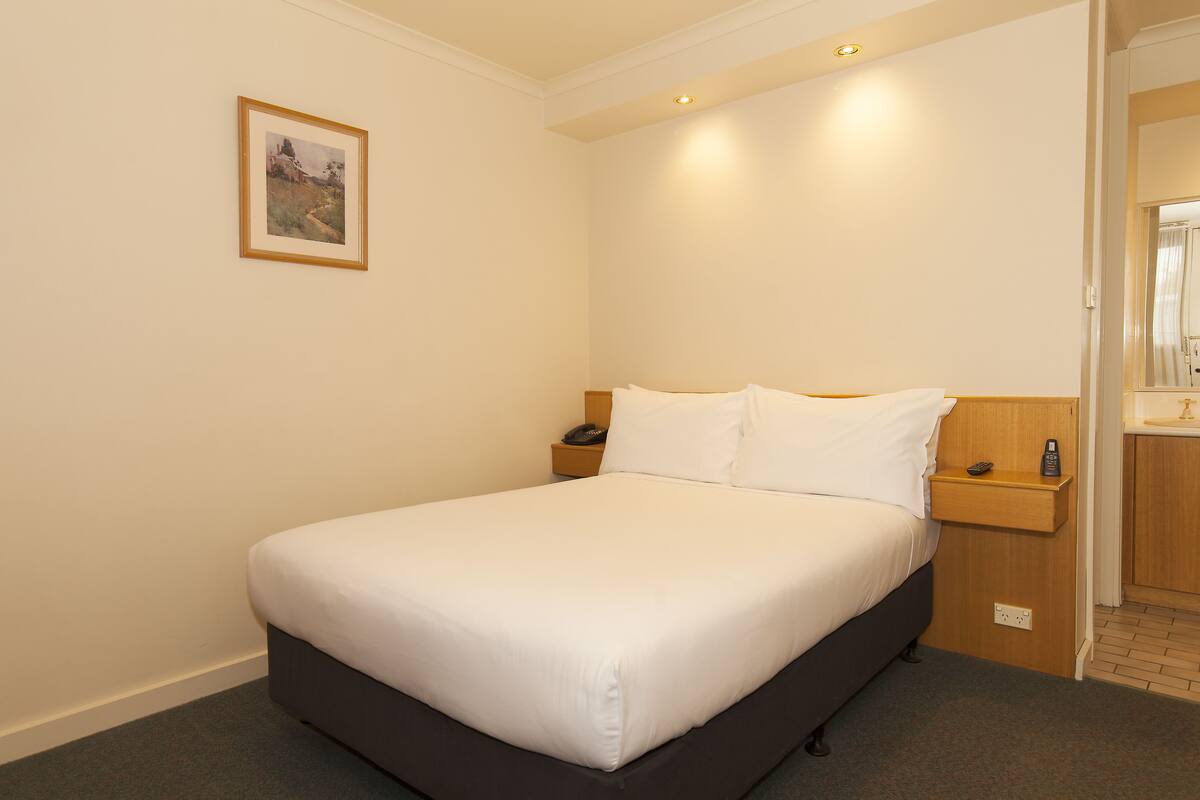 Cama bem arrumada com lençóis brancos, mesa de cabeceira com telefone e controle remoto, e um quadro na parede.