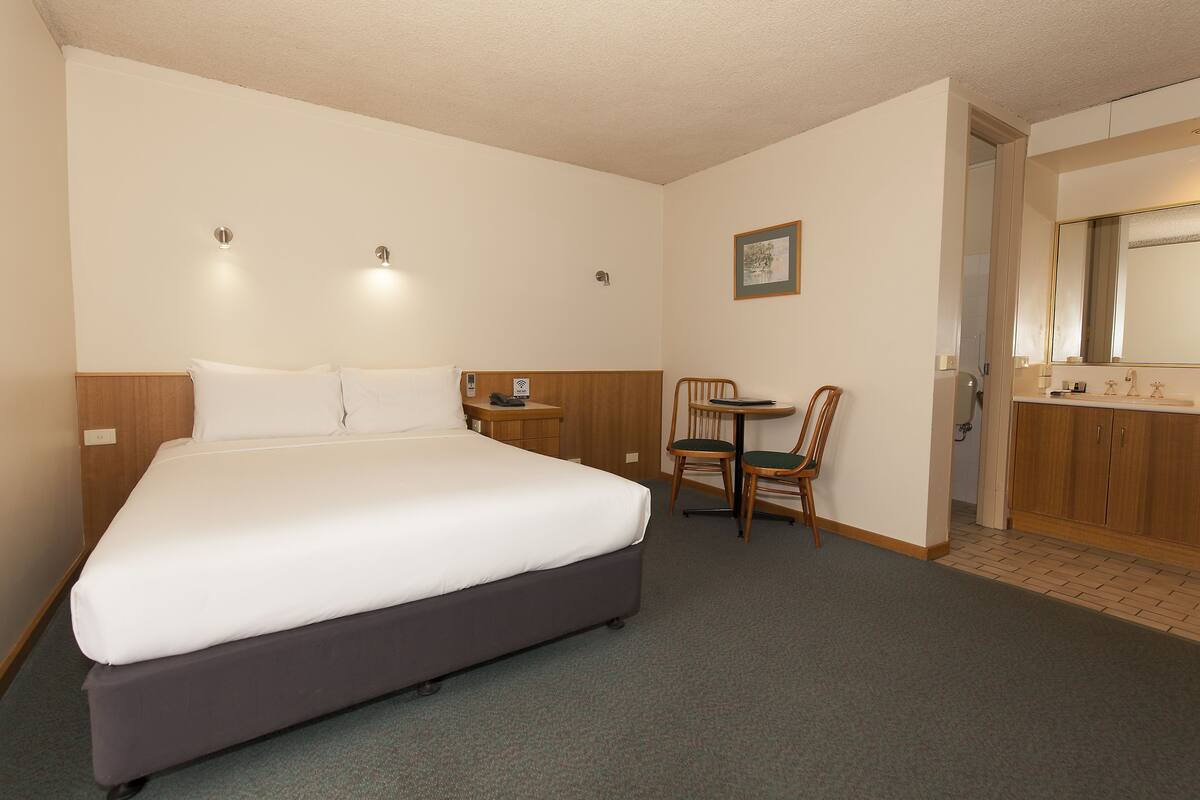 Quarto de hotel com uma cama grande, uma mesinha com cadeiras, banheiro com espelho e luminária fixada na parede.