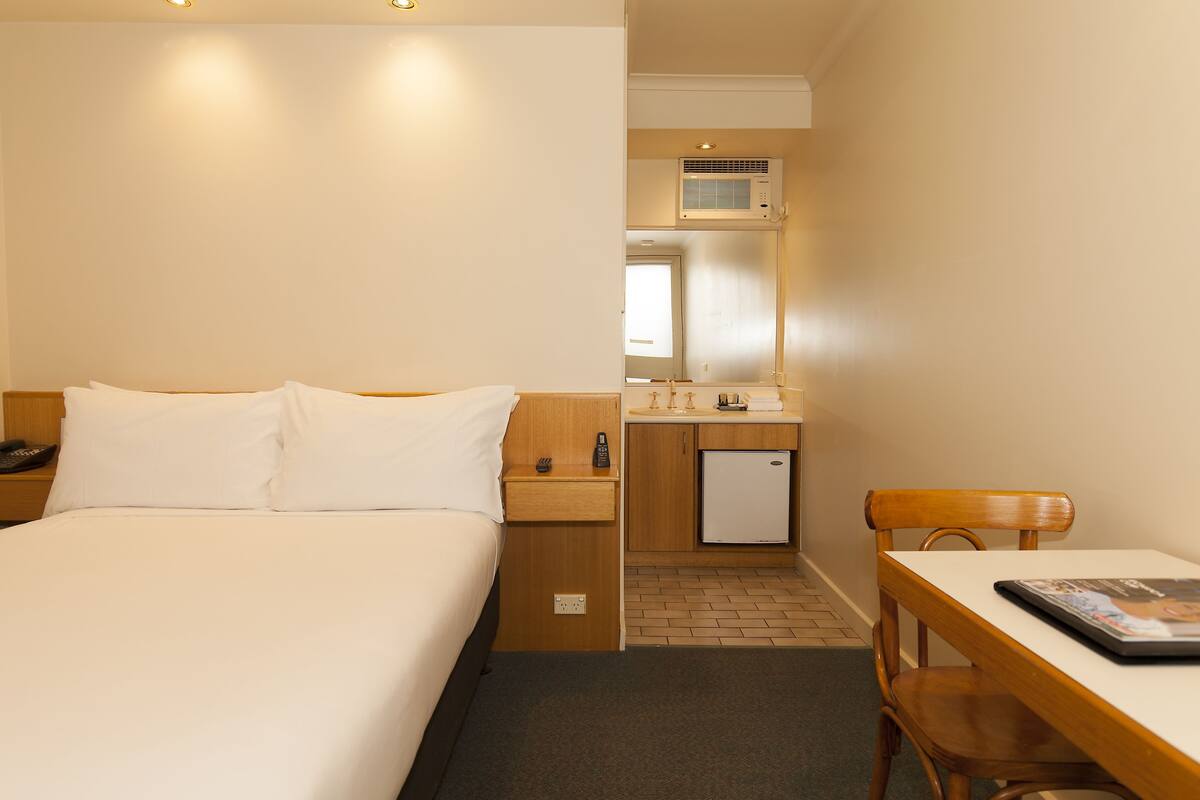 Quarto de hotel com cama, pequeno espaço de cozinha, espelho, mesa e cadeira.
