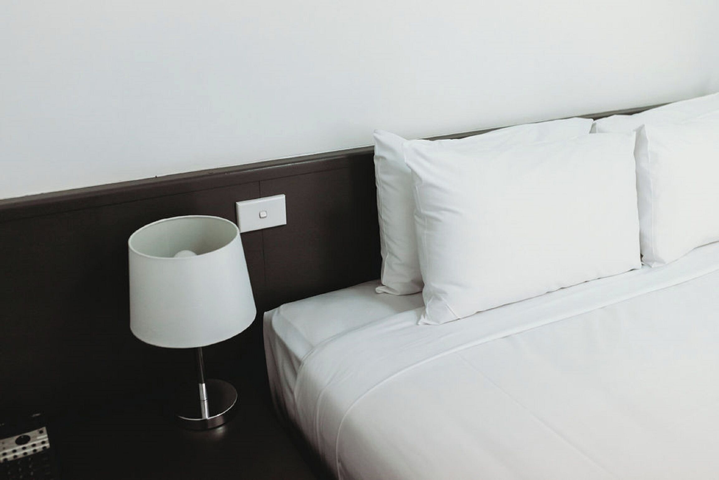 A neatly made bed with white bedding and pillows, a white bedside lamp, and a dark-colored nightstand.
