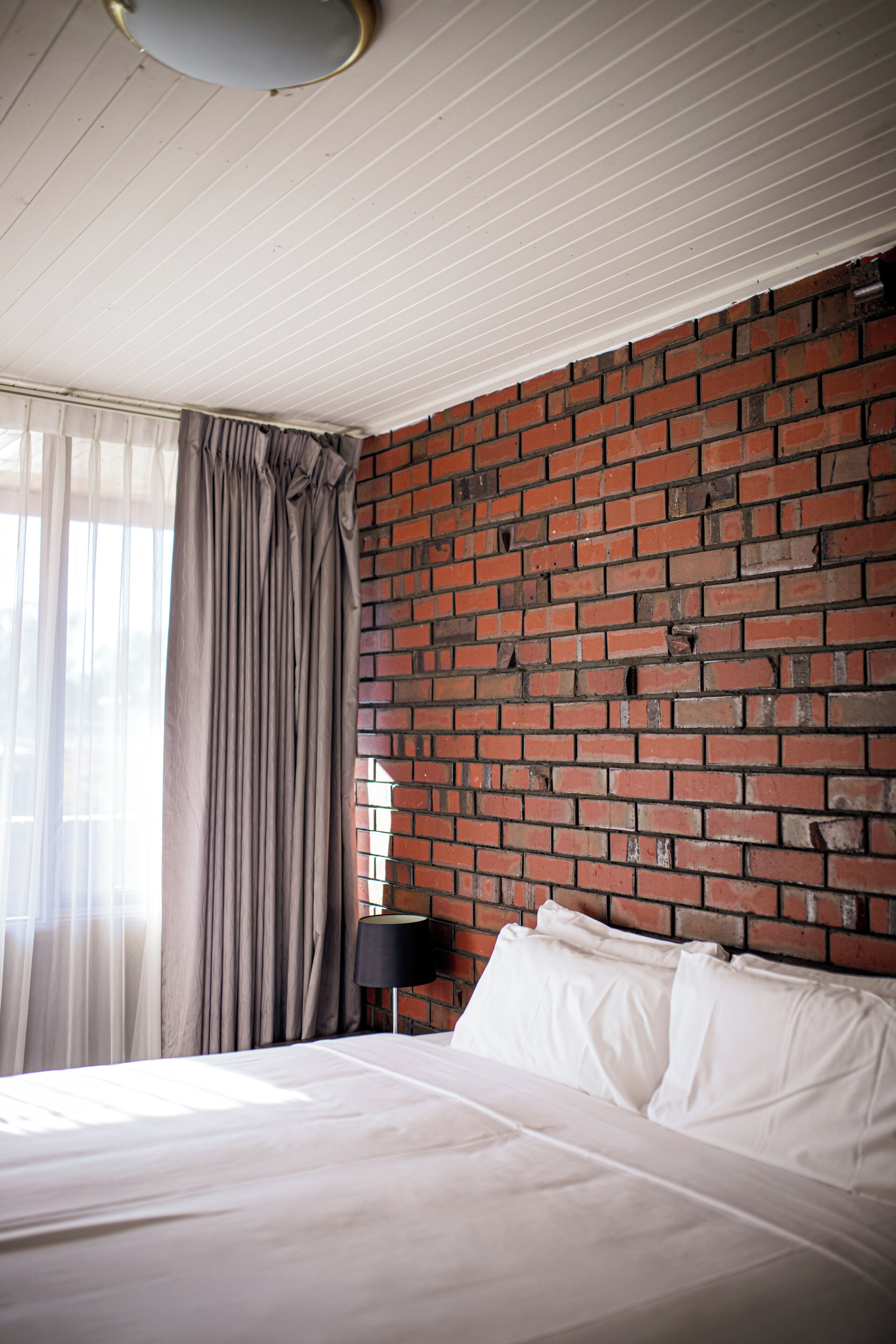 A hotel room with a brick wall, a bed with white linens, a lamp, and sheer curtains.