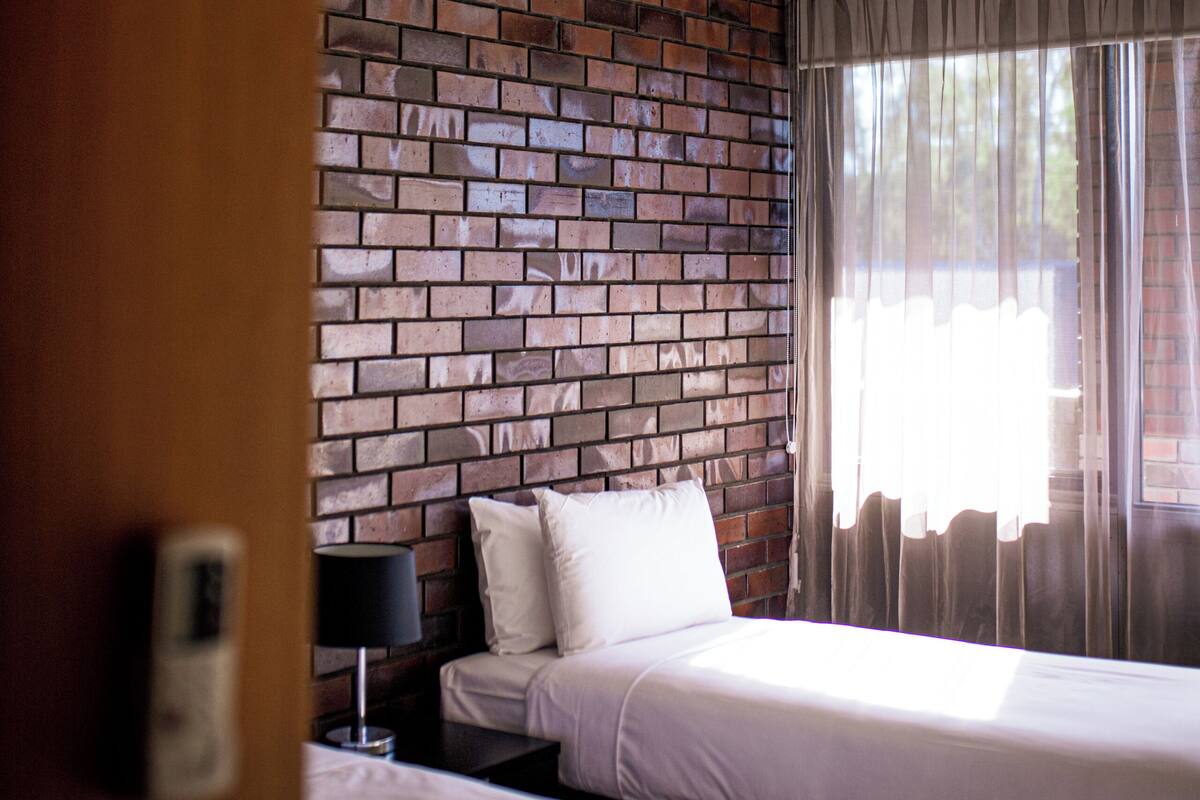 Quarto de hotel com duas camas, parede de tijolos, luminária no teto e cortinas finas.