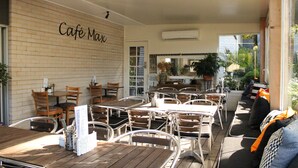An outdoor café with wooden tables and metal chairs, a brick wall with 'Café Max' signage, and a view of the street. - Prom Country Lodge (Foster)