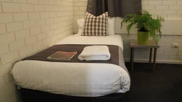 A neatly made bed with a checkered pillow, a folded white towel, and a book on the bed. There is a green potted plant on a small table beside the bed.