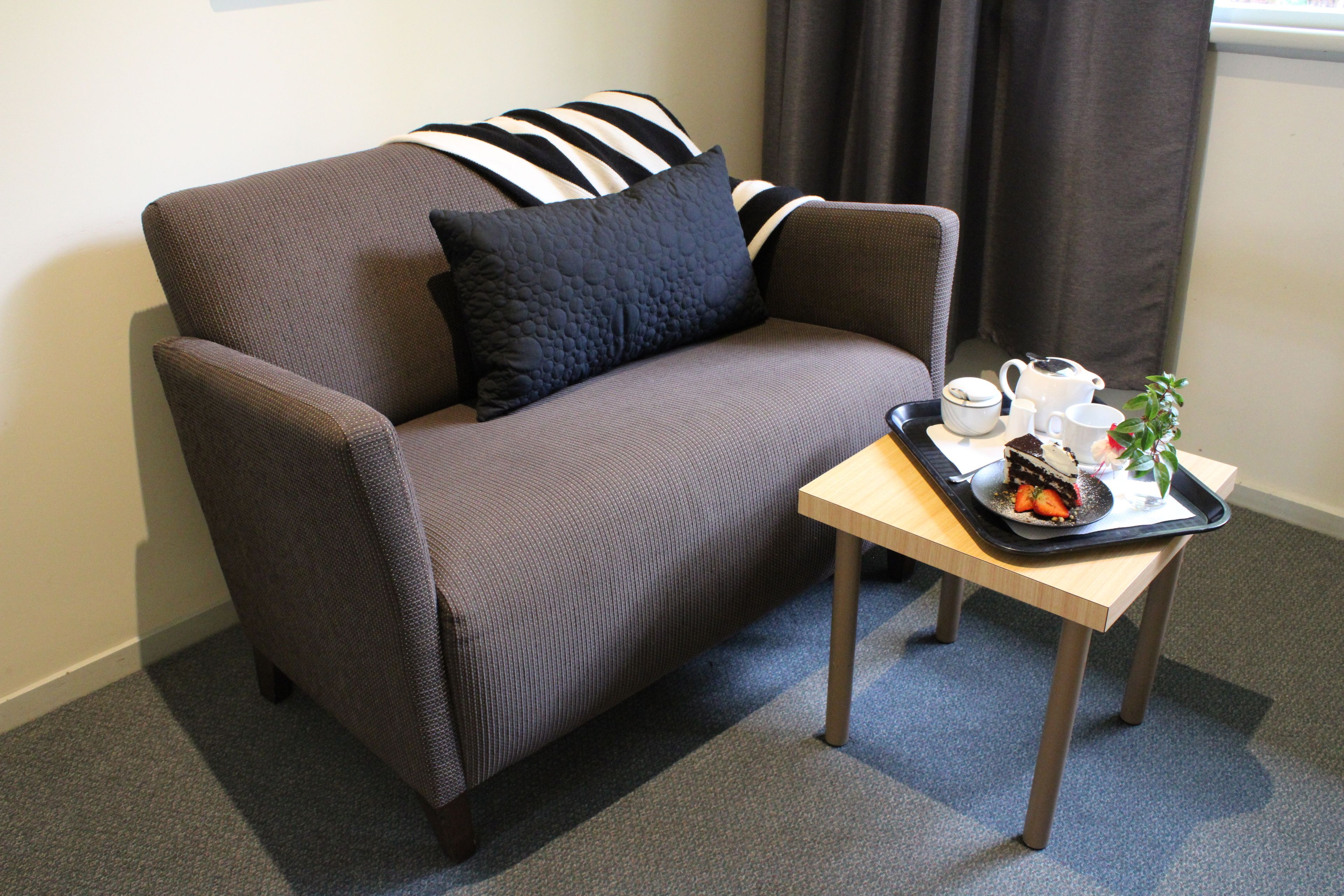 En brun soffa med en svart kudde och en randig kudde, ett brickbord med en kaka, en kopp och en växt på ett träbord.