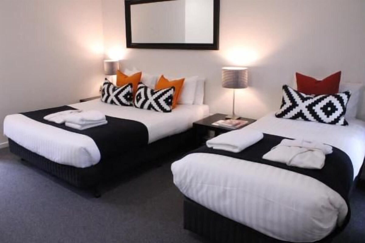 A hotel room with two beds, each with a black and white patterned pillow, a white blanket, and a red pillow. There is a nightstand with a lamp between the beds.