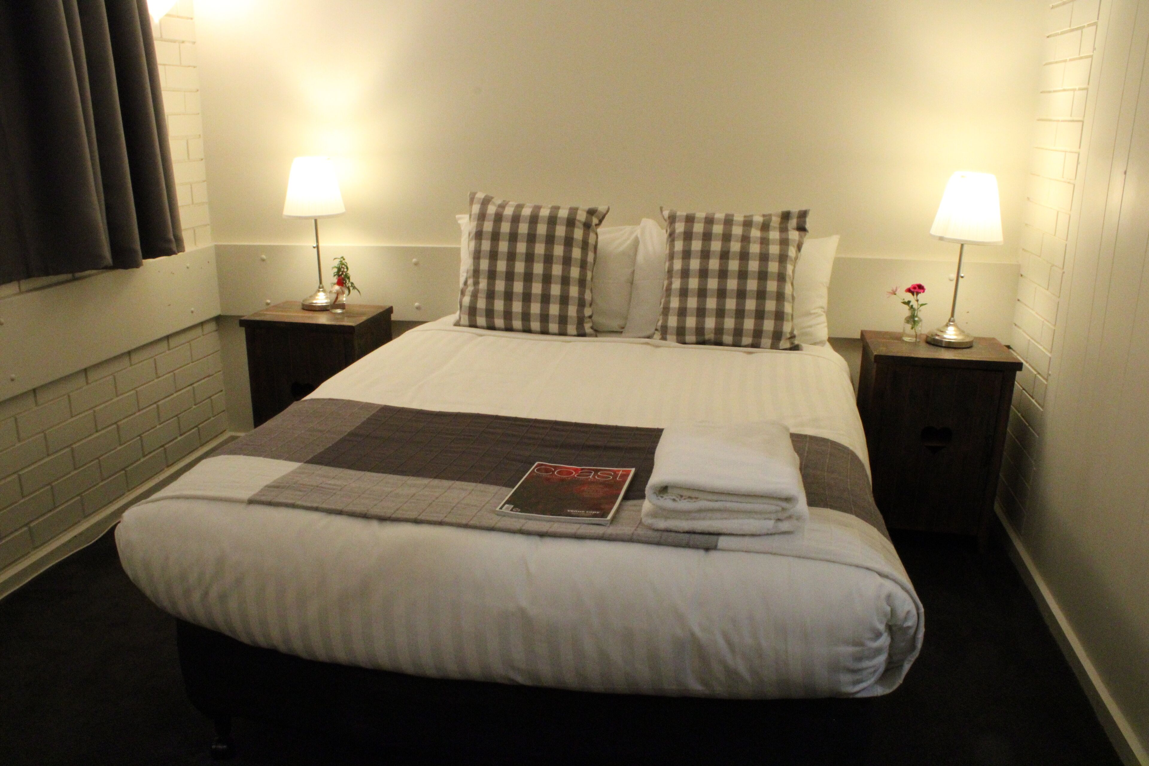 A neatly made bed with a checkered pillow set, a folded towel, and a magazine on the bed. There are two bedside tables with lamps and a small vase with flowers.