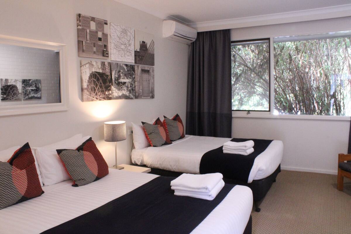A hotel room with two beds, a nightstand, a lamp, and a window with a view of trees.