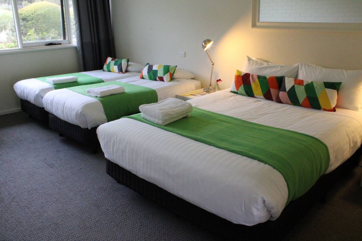 A hotel room with two single beds, each with a green bedspread and white linens, and a larger bed with a colorful geometric pillow.