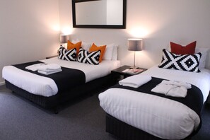 A hotel room with two beds, each with black and white bedding, orange and black patterned pillows, and white towels. There is a small table with a lamp between the beds.