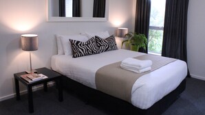 A neatly made bed with white and zebra-patterned pillows, a small bedside table with a magazine, and a potted plant. - Prom Country Lodge (Foster)