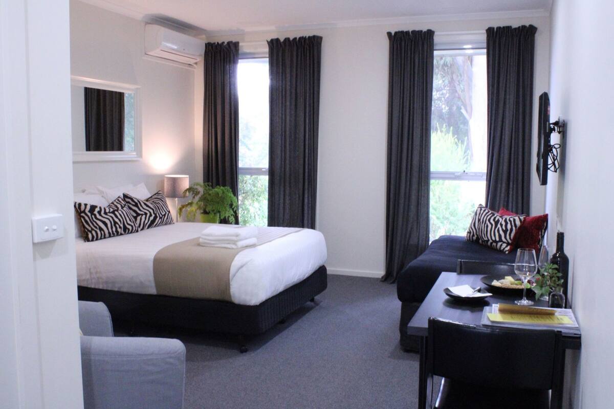 A hotel room with a bed, a sofa, a dining table with a chair, a potted plant, and a window with curtains.