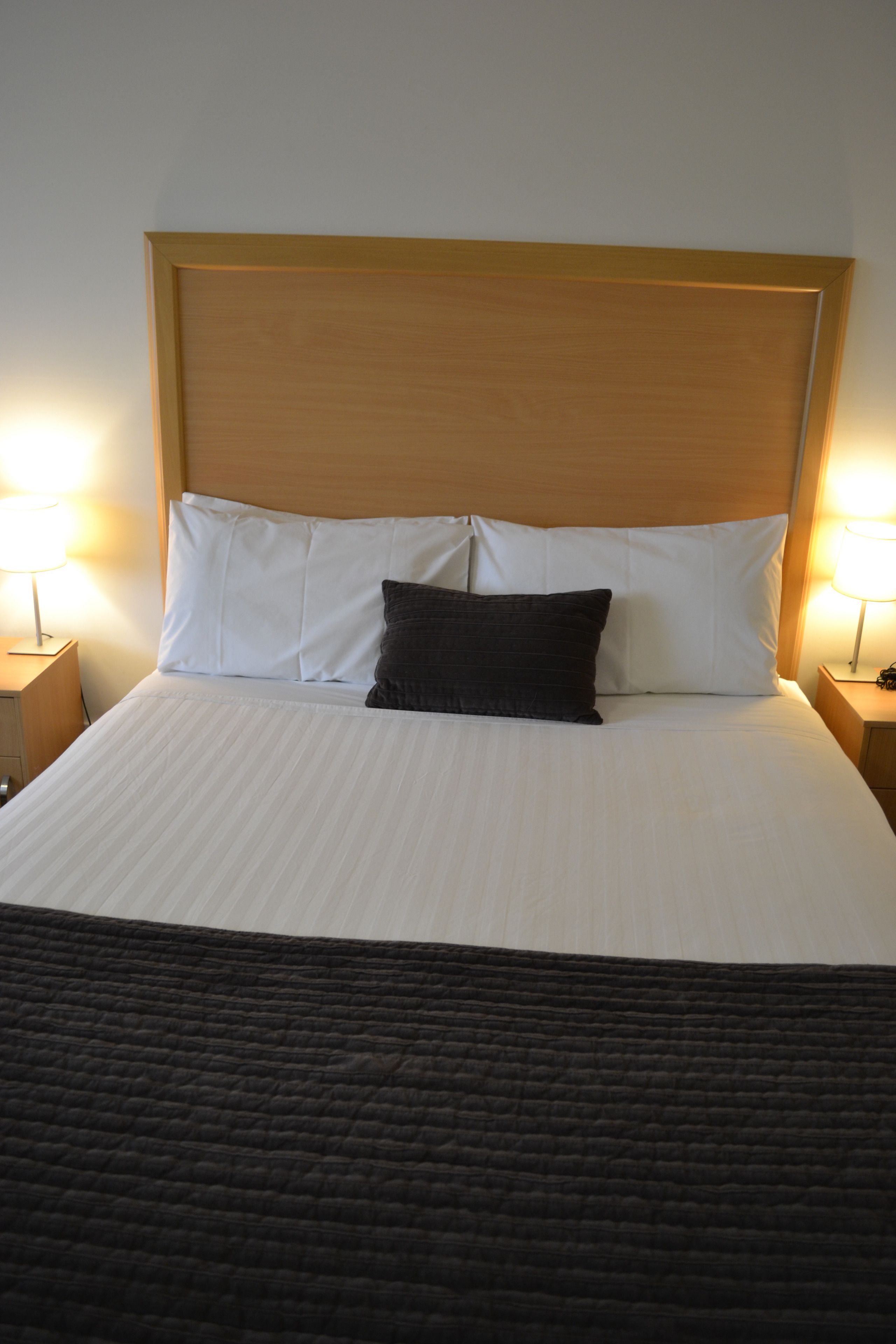 A neatly made bed with white linens and a dark brown pillow, flanked by two bedside lamps on wooden nightstands.
