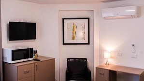 A hotel room with a flat-screen TV, a microwave, a framed abstract painting, a black leather chair, a bedside table with a lamp, and a wooden cabinet.