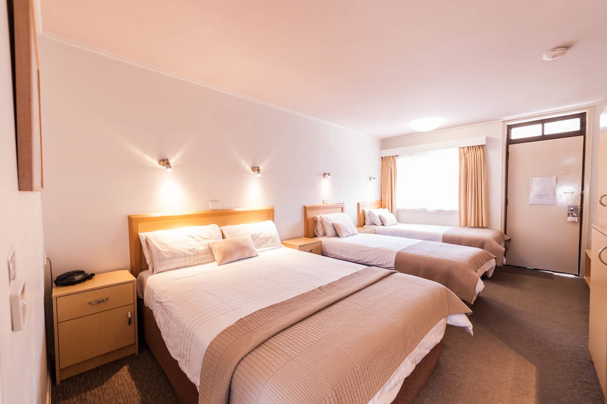 A hotel room with three beds, a wooden nightstand, a desk, and a door with a window and curtains.