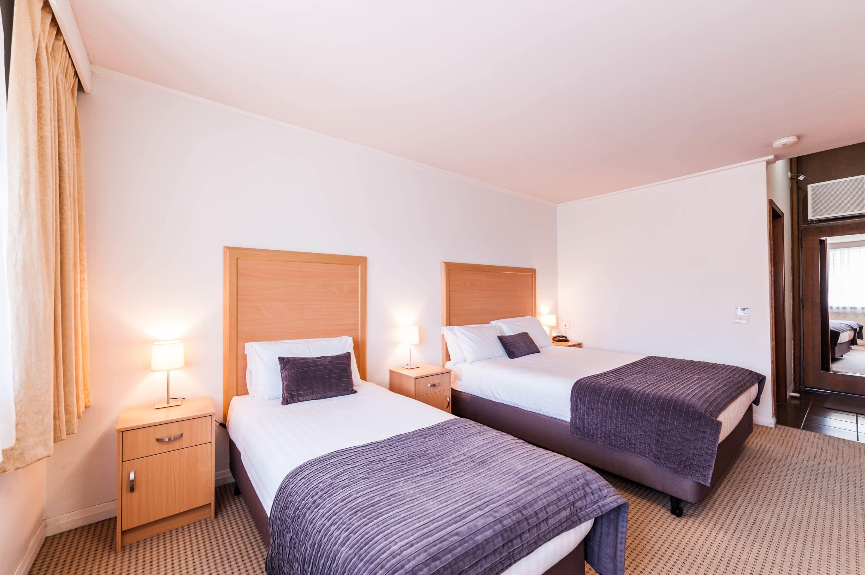 Une chambre d’hôtel avec deux lits, des têtes de lit en bois, des tables de chevet avec des lampes et un sol recouvert de moquette.