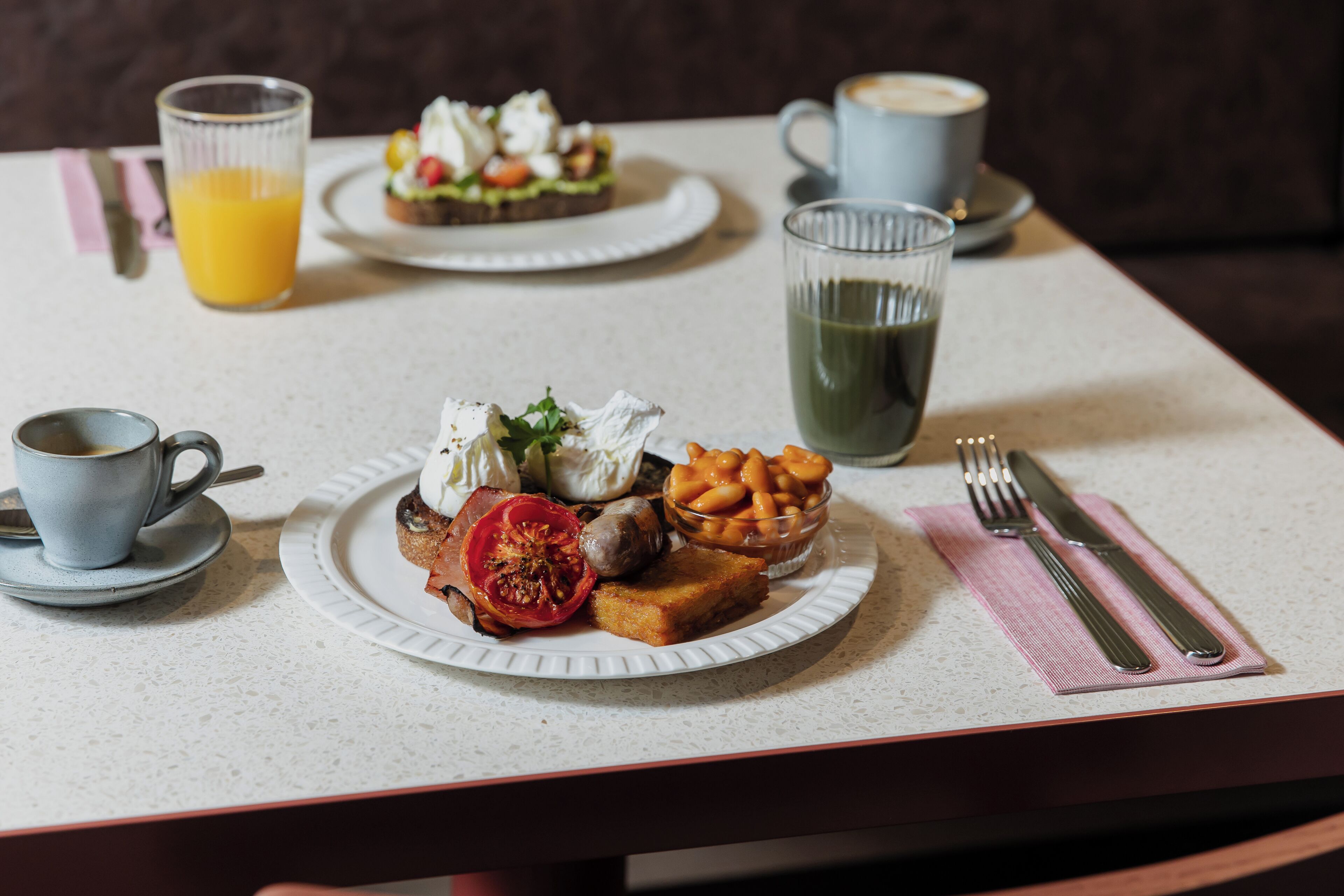 Une table dressée pour le petit-déjeuner avec une assiette de toasts, des œufs, des tomates et des haricots, accompagnée d’un verre de jus d’orange et d’une tasse de café.