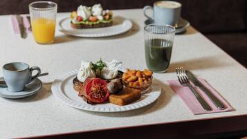 Une table dressée pour le petit-déjeuner avec une assiette de toasts, des œufs, des tomates et des haricots, accompagnée d’un verre de jus d’orange et d’une tasse de café.