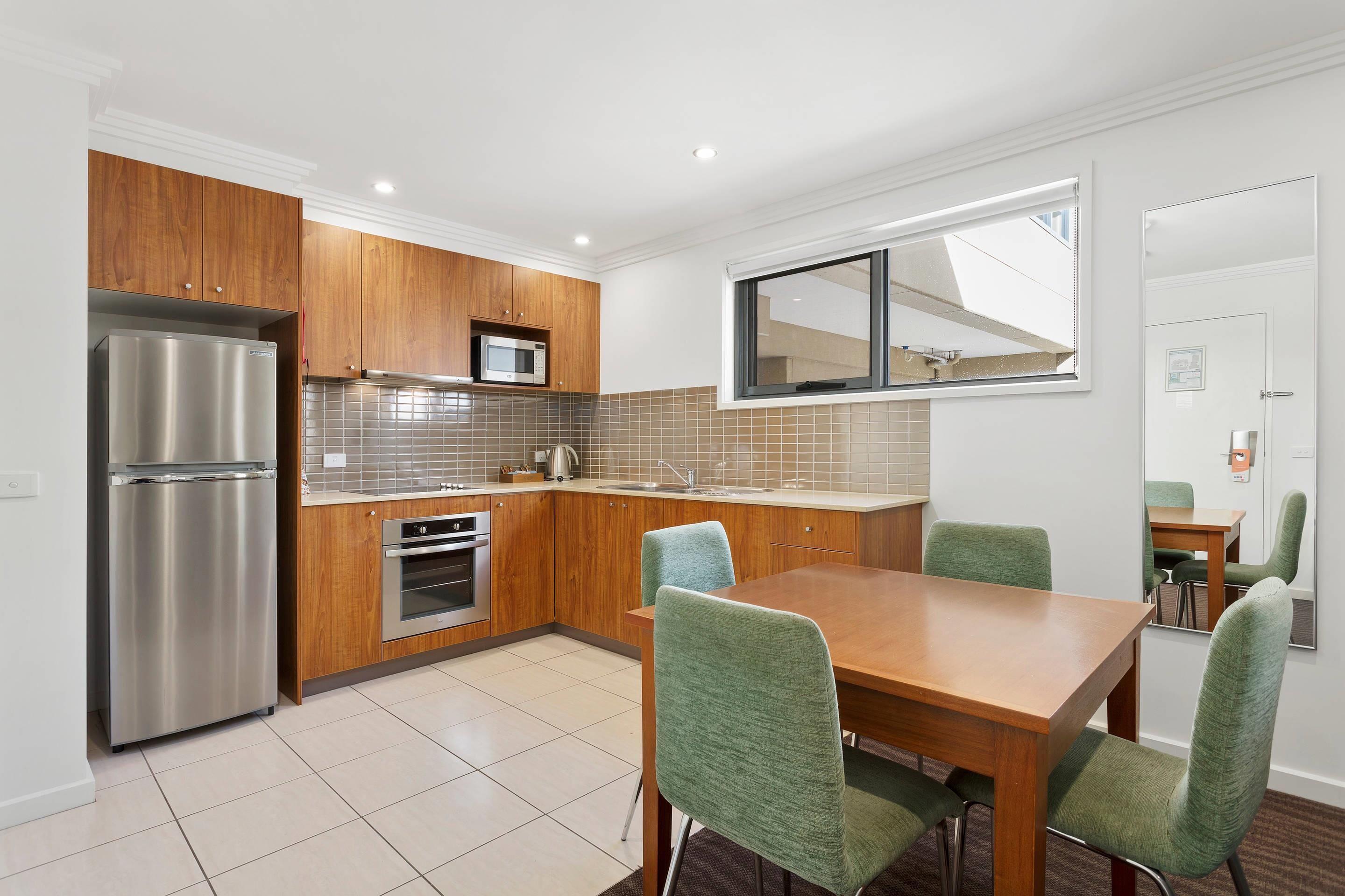 A modern kitchen with wooden cabinets, stainless steel appliances, a dining table with chairs, and a window with blinds.