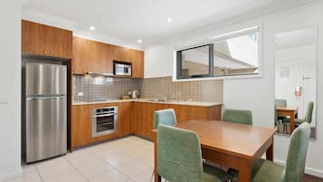 A modern kitchen with wooden cabinets, stainless steel appliances, a dining table with chairs, and a window with blinds.