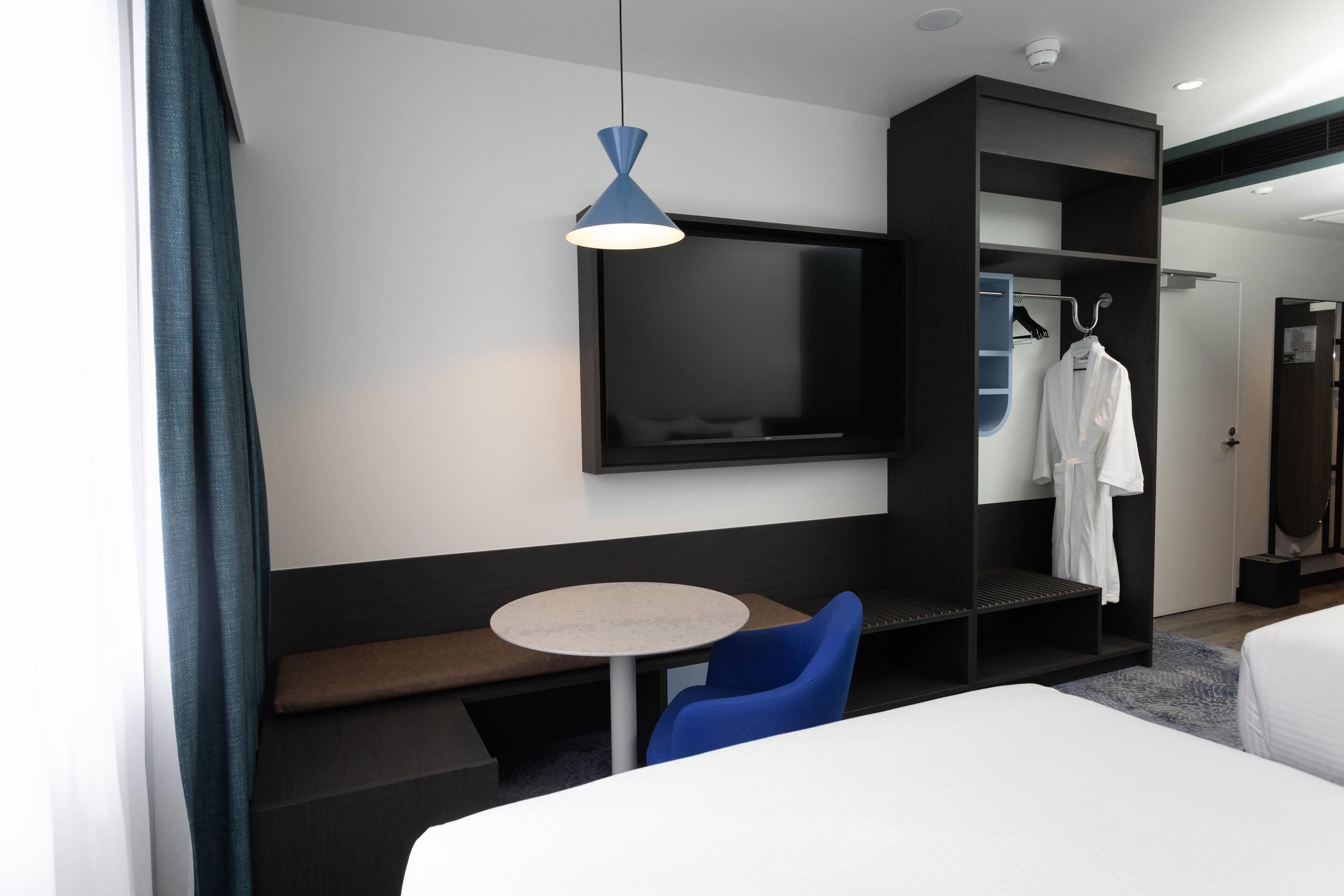 Habitación de hotel con cama, una mesa redonda, una silla azul, un televisor montado en la pared y un armario con una bata colgando en su interior.