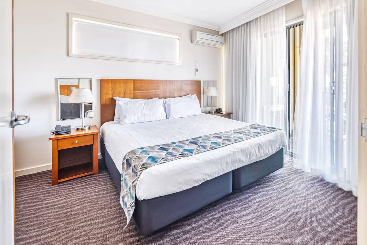 Hotelkamer met een groot bed, nachtkastje met lamp, spiegel en raam met dunne gordijnen.