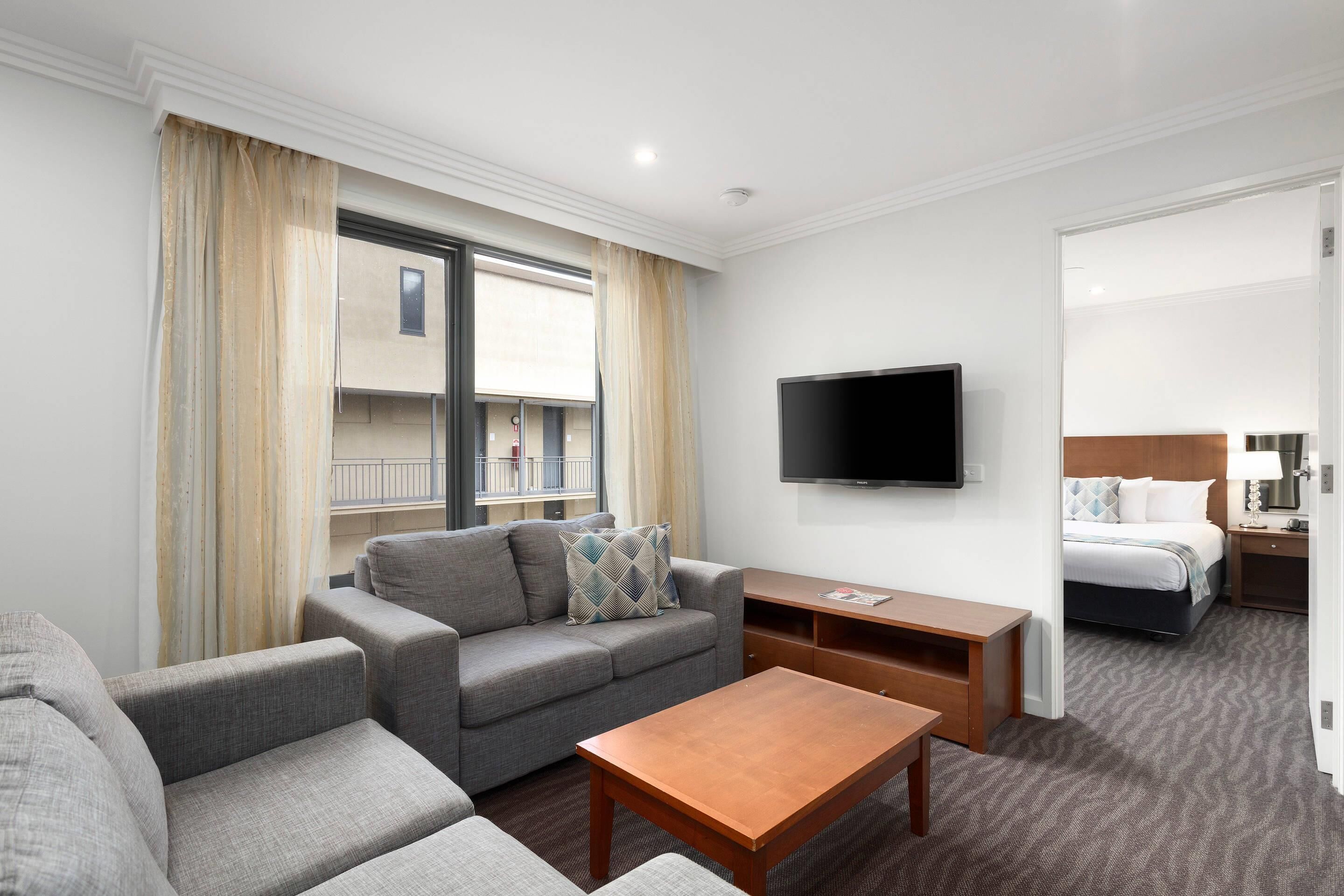 A hotel room with a sofa, a coffee table, a TV, a bed, and a window with curtains.