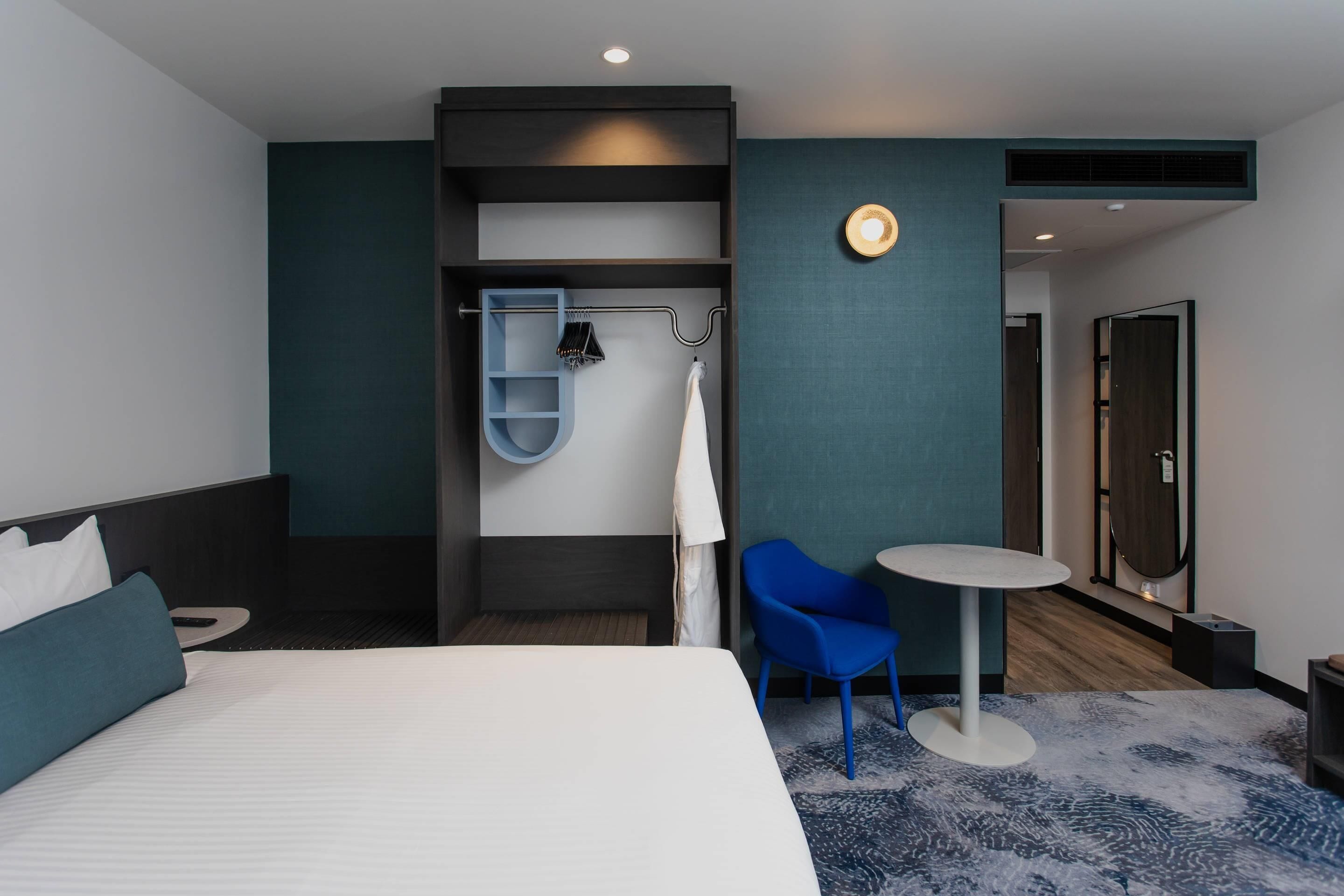 A hotel room with a bed, a seating area with a table, a coat rack, and a mirror.