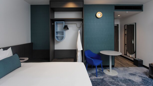 Ein modernes Hotelzimmer mit einem großen Bett, einem kleinen runden Tisch, einem blauen Sessel, einer eingebauten Ablage und einem Spiegel.