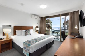 Habitación de hotel con cama, mesitas de noche, escritorio, silla, televisor y balcón con vista al mar.