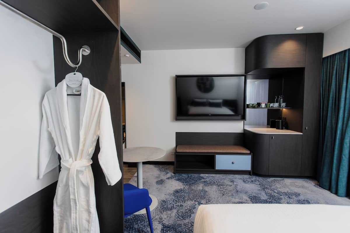 Een moderne hotelkamer met een bed, een televisie, een tafeltje, een bankje, een badjas en een wastafel.