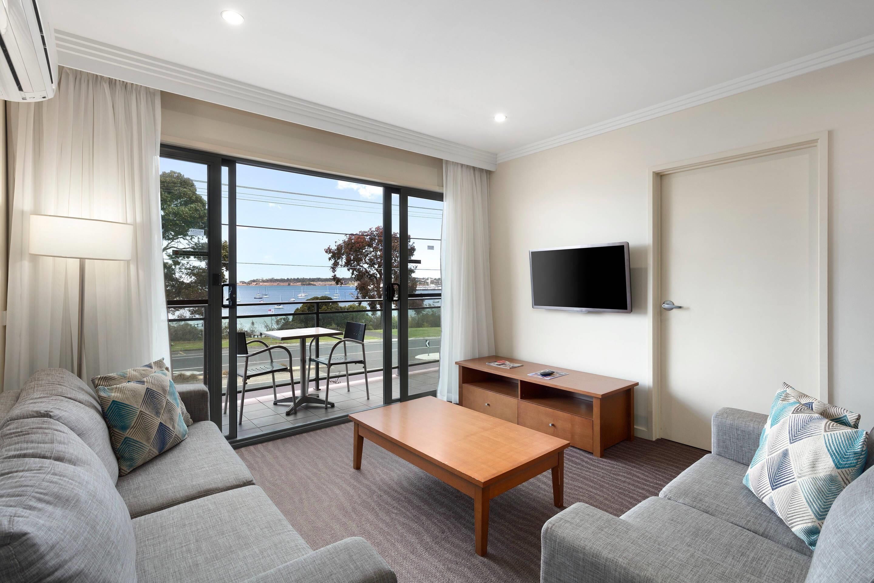 A modern hotel room with a sofa, a wooden coffee table, a TV mounted on the wall, and a balcony with a view of the sea.