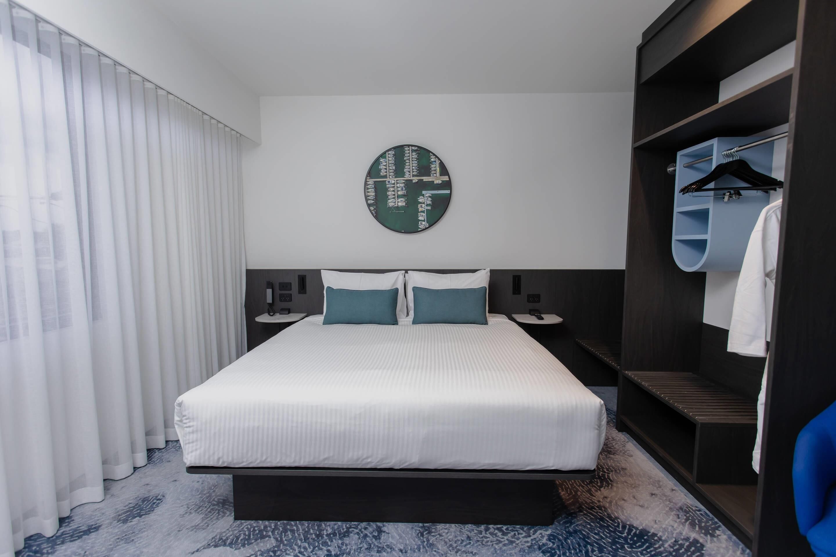 A modern hotel room with a large bed, a round wall clock, a wardrobe, and a mirror.