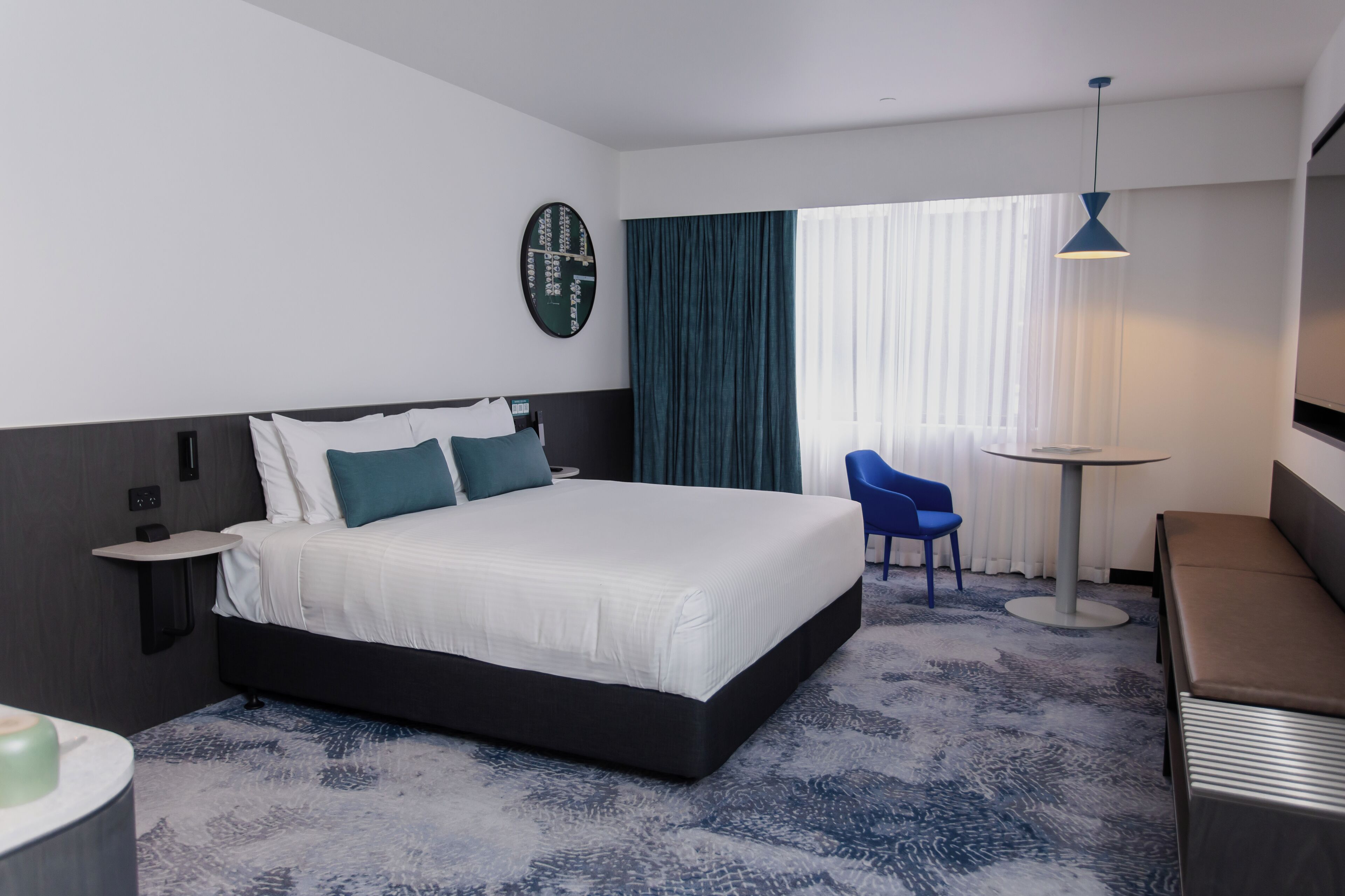 A modern hotel room with a large bed, a bedside table, a small round table, a blue chair, and a sofa.
