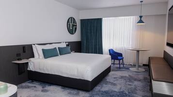 A modern hotel room with a large bed, a bedside table, a small round table, a blue chair, and a sofa.
