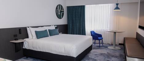 A modern hotel room with a large bed, a bedside table, a small round table, a blue chair, and a sofa.