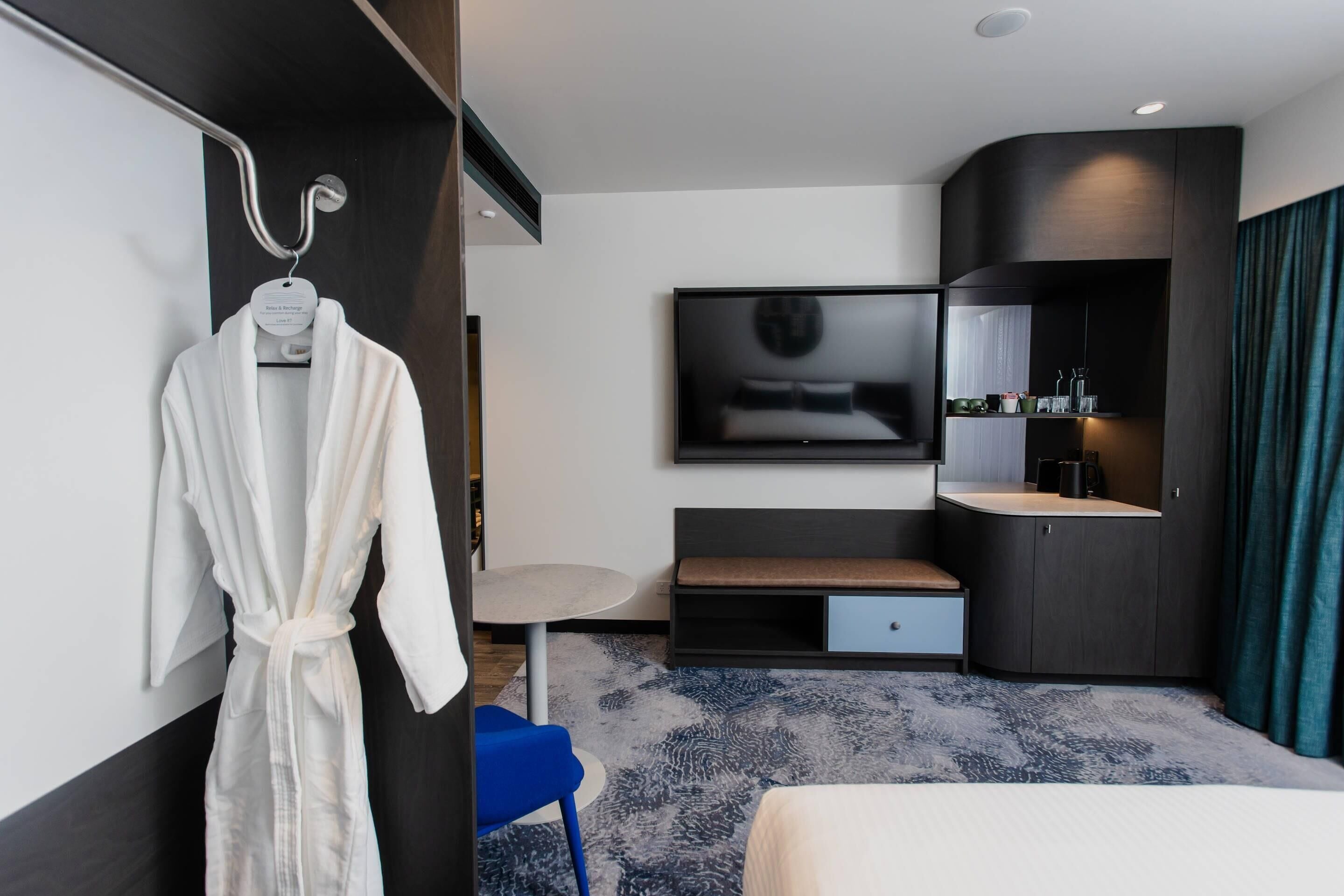 A modern hotel room with a bed, a TV, a small table, a bench, a robe, and a bathroom area.