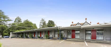 Fila de habitaciones de motel con puertas rojas, un estacionamiento y algo de vegetación al frente.