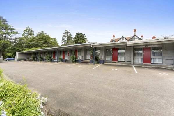 A row of motel rooms with red doors, a parking lot, and some greenery in the foreground.