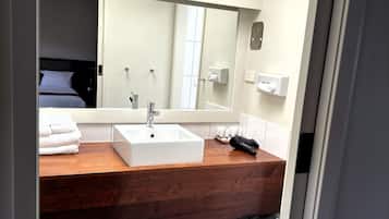 Une salle de bain moderne avec un grand miroir, un lavabo blanc, un plan de travail en bois et une poubelle visible.