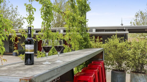 A bar area with a bottle of wine and glasses on a wooden ledge, surrounded by red stools and greenery. - Botanical Motel (Hamilton)
