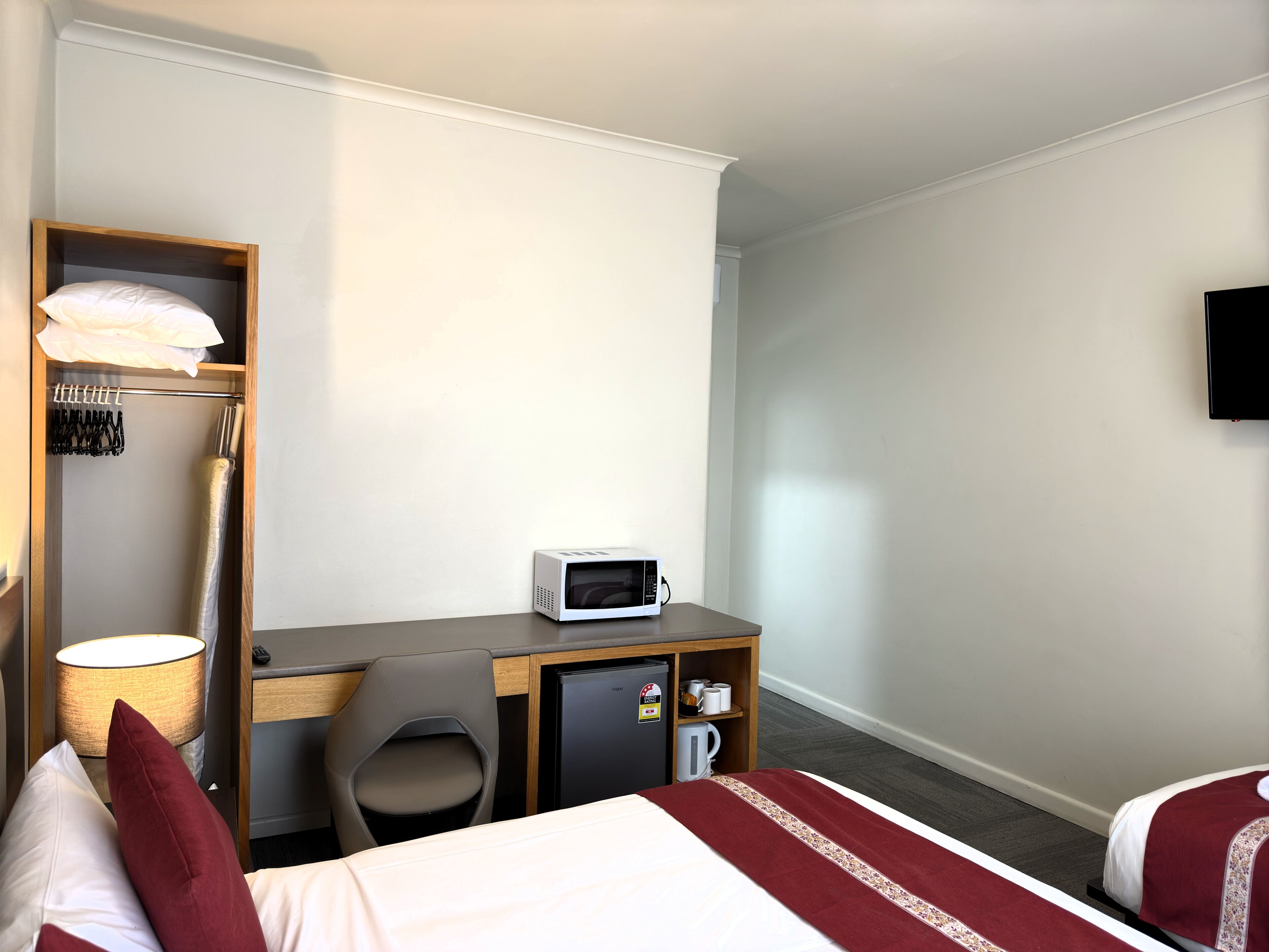 Habitación de hotel con una cama, un escritorio con microondas, una silla, un refrigerador pequeño, una lámpara y un televisor en la pared.