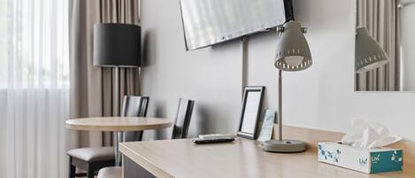 A hotel room with a desk, chairs, a lamp, a TV, a mirror, and a tissue box.