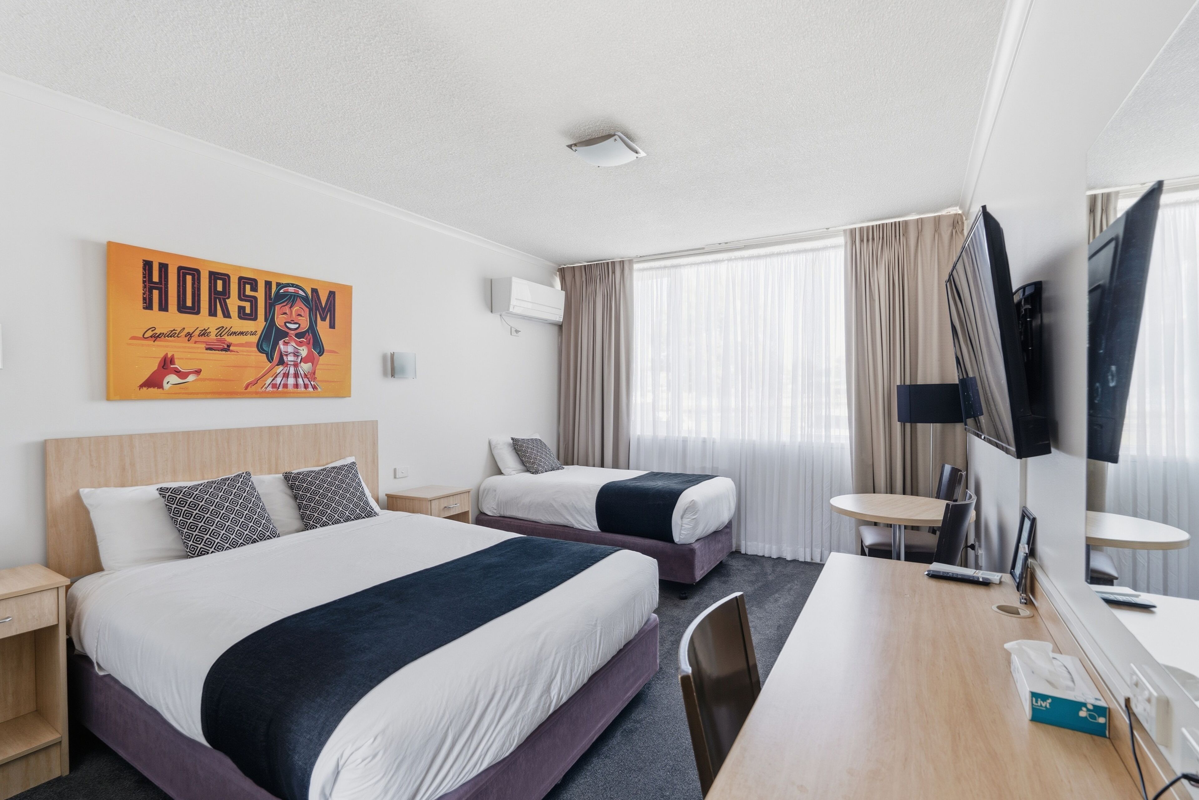 A hotel room with two beds, a desk, a TV, and a large window with curtains.
