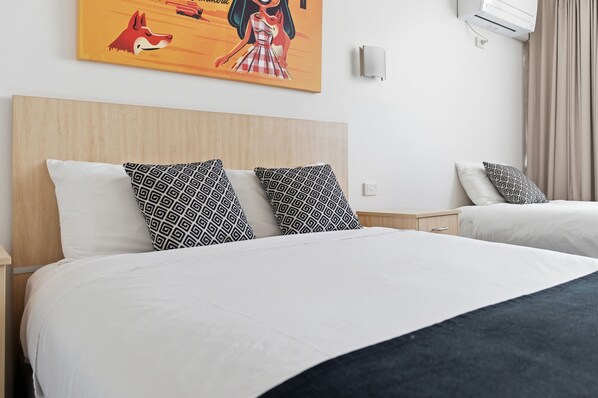 A hotel room with two beds, patterned pillows, a headboard, a wall-mounted air conditioner, and a painting.