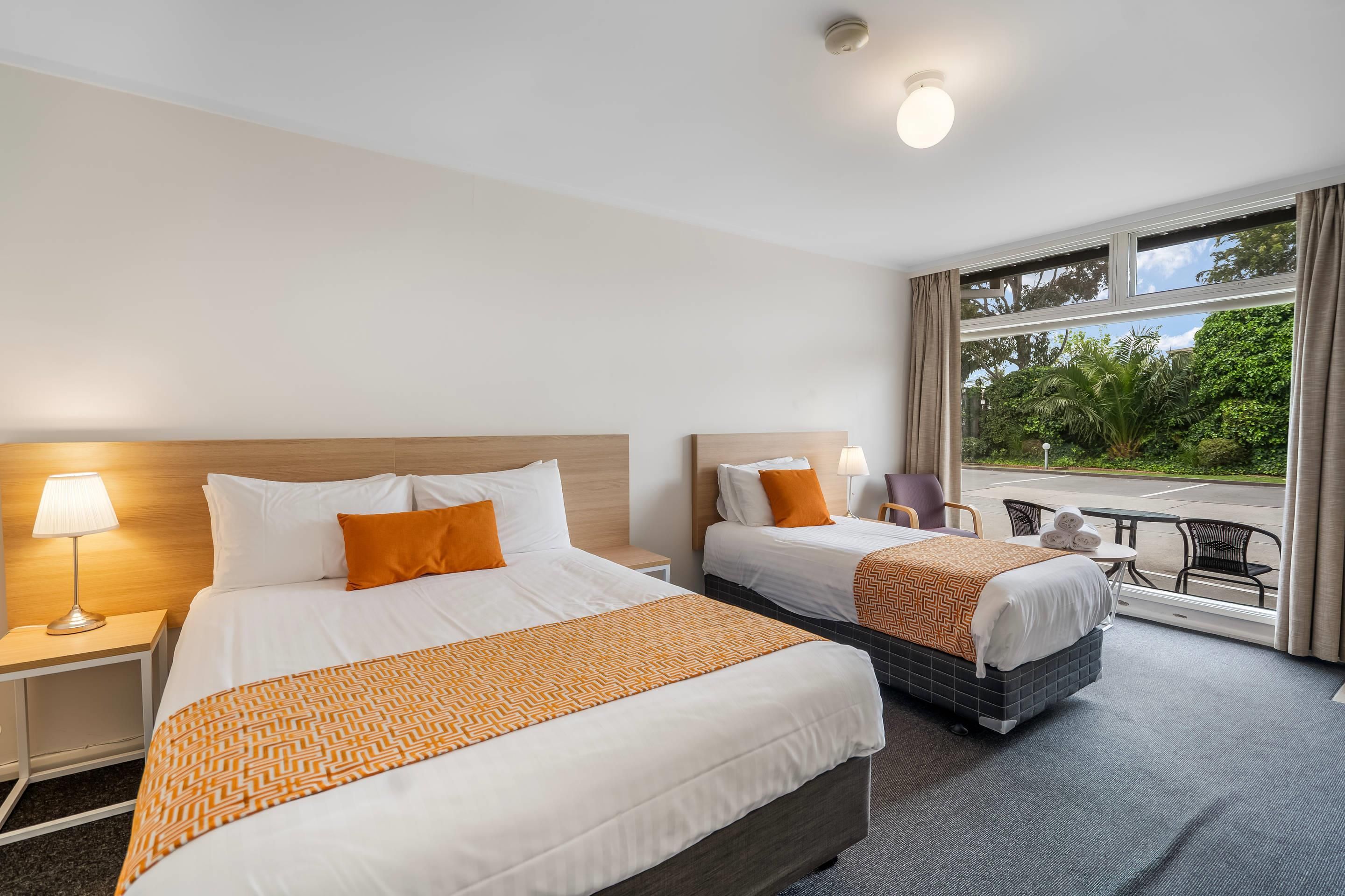 A hotel room with two beds, a small table, chairs, and a view of the outdoors.