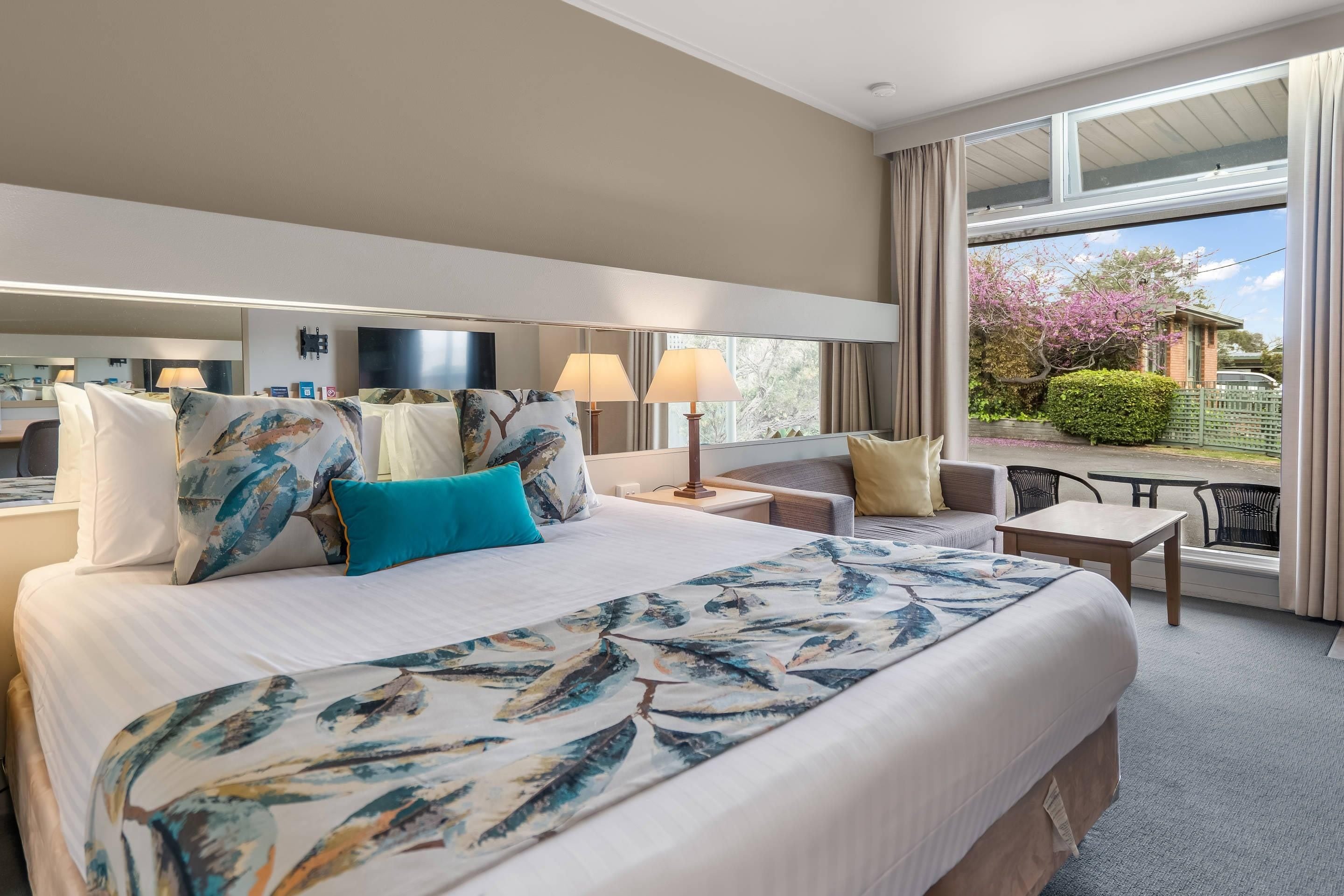 Habitación de hotel con una cama grande, dos mesitas de noche, una zona de estar con una silla y una pequeña mesa, y un balcón con vistas al exterior.