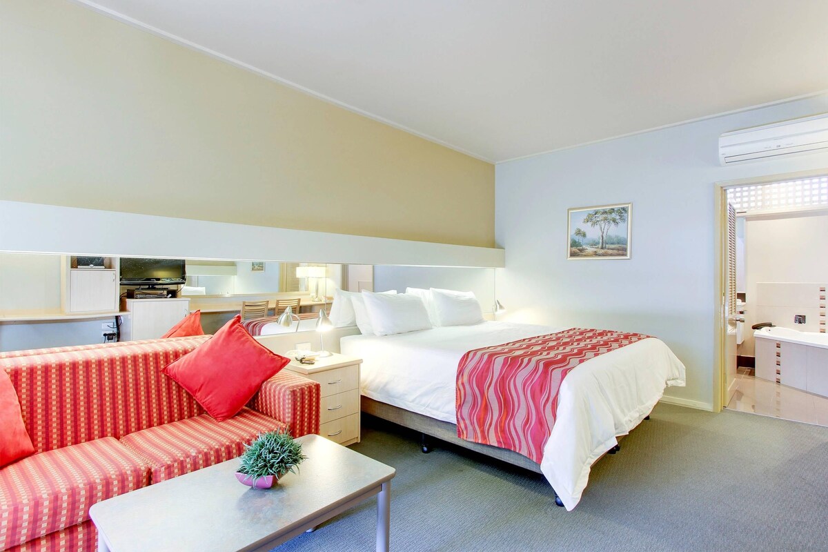 A hotel room with a bed, a sofa, a small table with a plant, a mirror, and a picture on the wall.